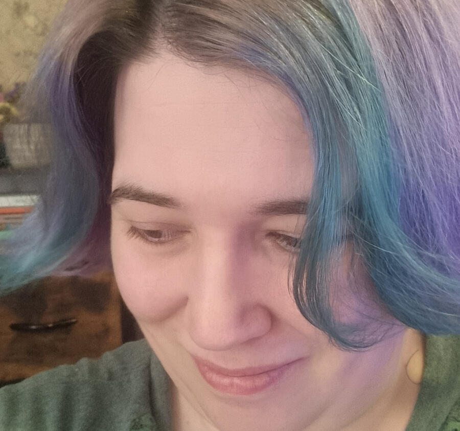 Rebecca, a white woman with short purple and teal hair, looks down and to the left with a small smile on her face.