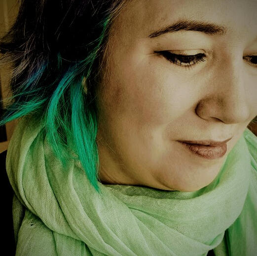 Rebecca, a white woman with short purple and blue hair, looks to the side. She is wearing a light green scarf.